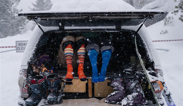 Two people showing their ski socks sitting in the back of a car with the trunk open and snow on the ground.